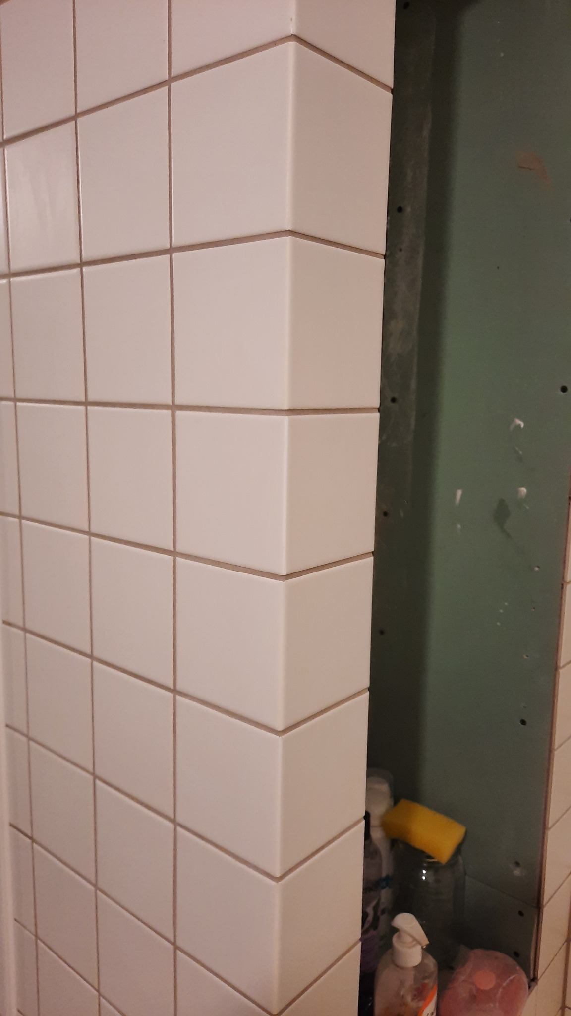 Biała, kwadratowa glazura ułożona na rogu ściany, z widocznymi fugami w kolorze beżowym, obok surowa ściana w kolorze zielonym z widocznymi otworami montażowymi i fragmentem półki z akcesoriami...