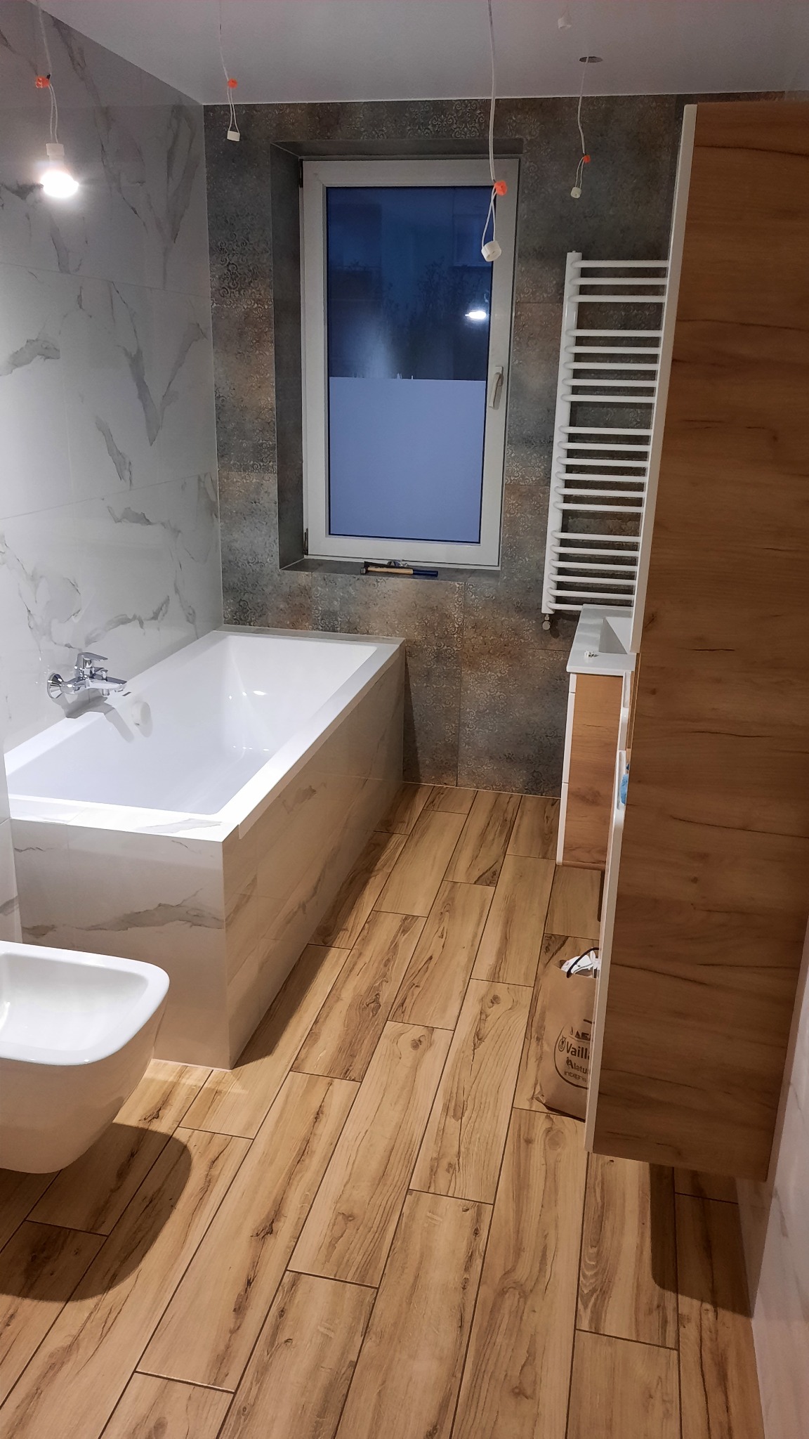 Nowoczesna łazienka z wanną obudowaną płytkami imitującymi marmur, drewnopodobną podłogą, grzejnikiem drabinkowym i częściowo zamontowanym oświetleniem sufitowym.