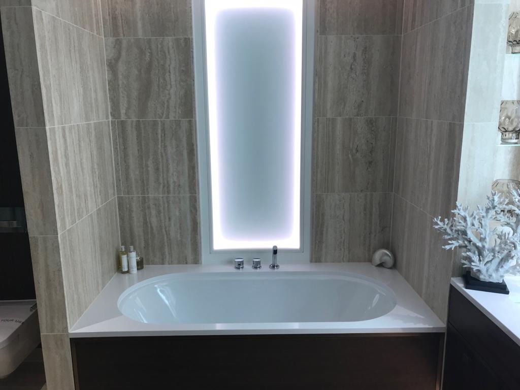 Nowoczesna łazienka z wanną obudowaną ciemnym drewnem, ściany wyłożone płytkami imitującymi trawertyn, podświetlane pionowe okno i dekoracja w kształcie koralowca.