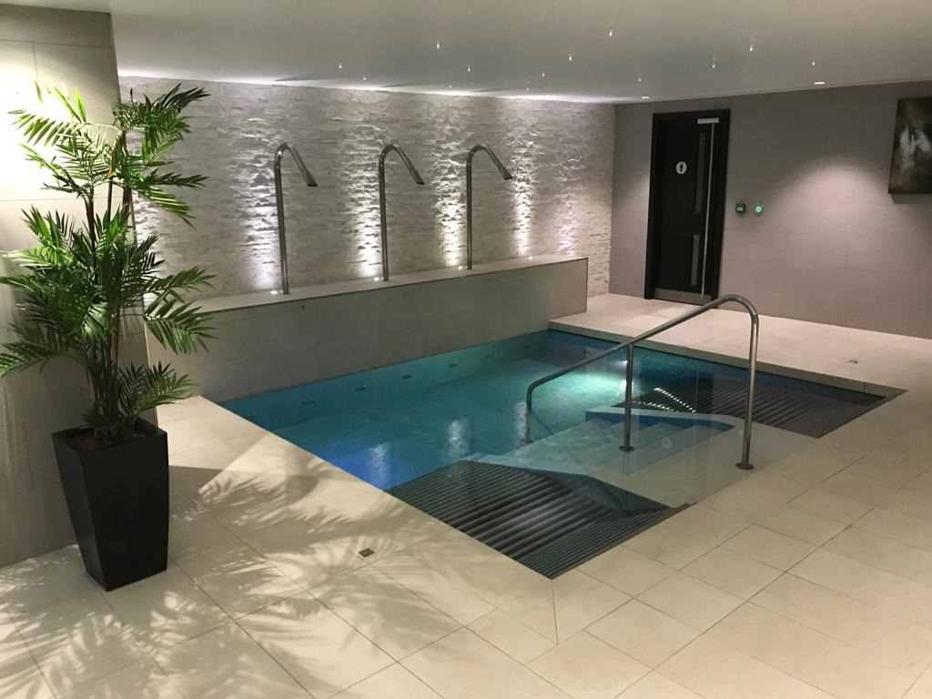 Kryty basen z turkusową wodą, schodami wejściowymi i metalową barierką, otoczony białymi płytkami, z dekoracyjną ścianą z białej cegły i rośliną w czarnej donicy.