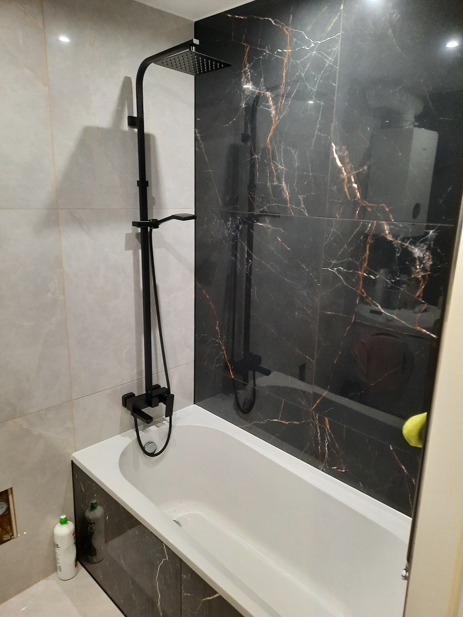 Nowoczesna łazienka z czarną armaturą i marmurowymi płytkami w odcieniach szarości i czerni, widoczna biała wanna z obudową z płytek marmurowych, butelki z płynami na podłodze w rogu.