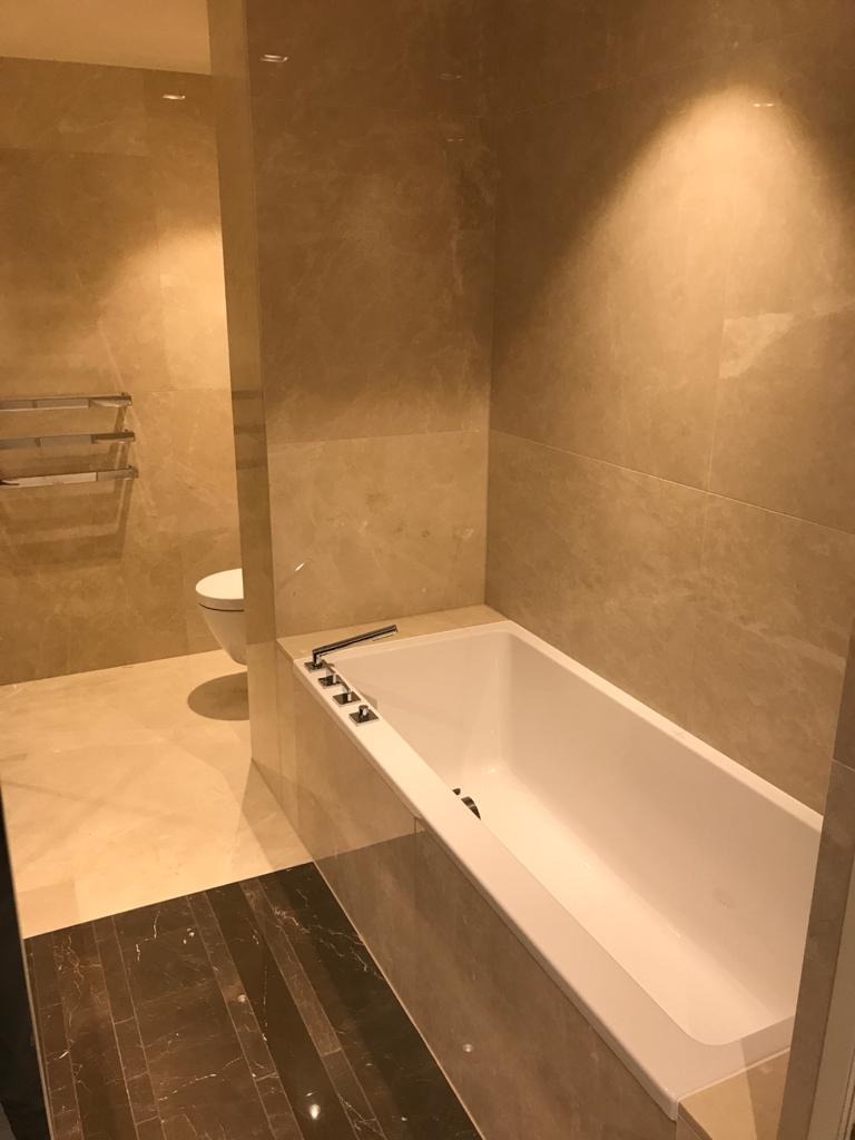 Wykończona łazienka z beżowymi marmurowymi płytkami na ścianach i podłodze, biała wanna z chromowaną armaturą, widoczny fragment toalety i ciemnobrązowa marmurowa podłoga przy wejściu.