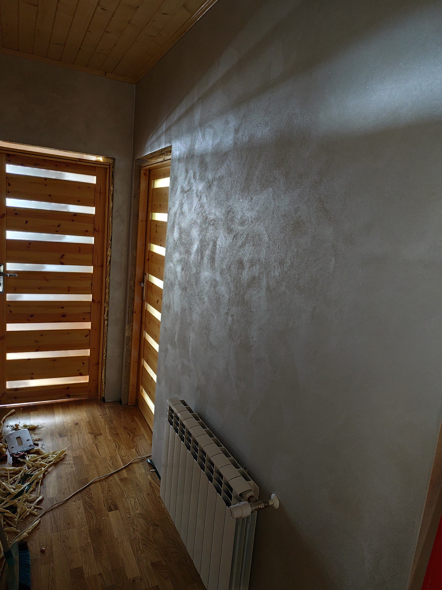 Ściana z efektem dekoracyjnym w odcieniach szarości, drewniany sufit i drzwi z poziomymi szczeblami. Widoczny grzejnik i fragment podłogi z resztkami materiałów budowlanych.