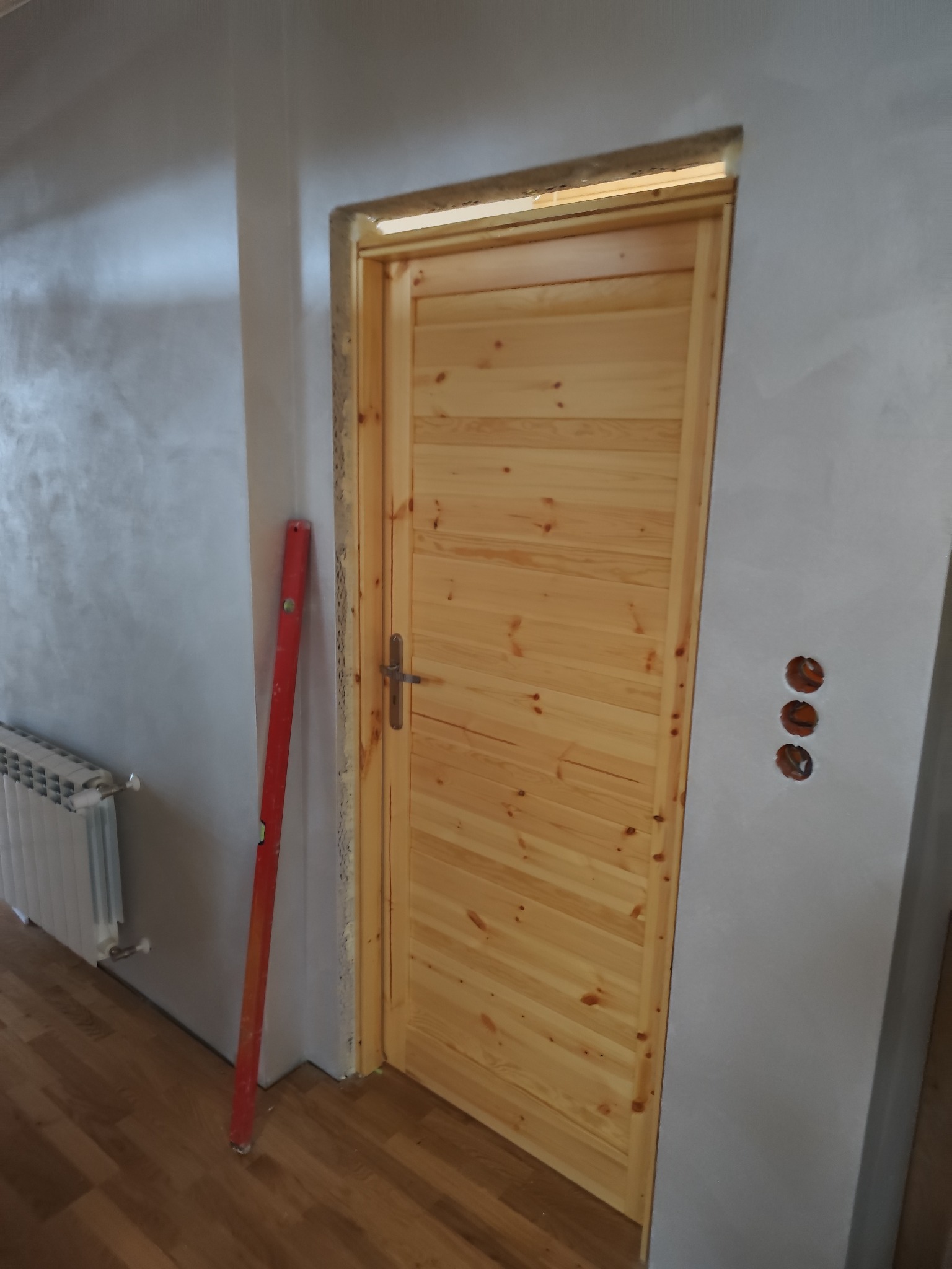 Jasne, drewniane drzwi z poziomym wzorem desek, zamontowane w szarej ścianie. Obok poziomica i widoczne puszki elektryczne. Fragment grzejnika w lewym dolnym rogu.