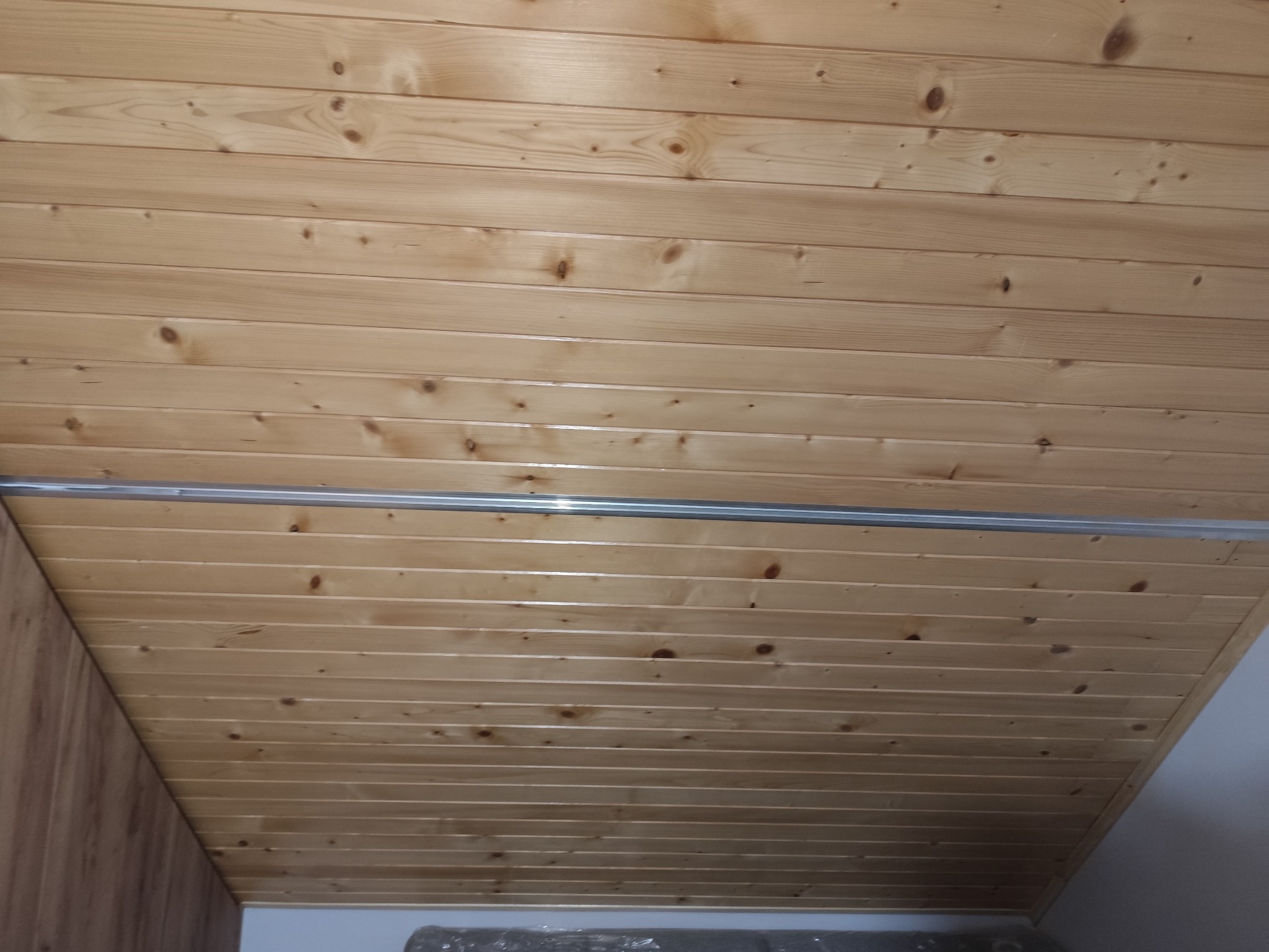 Drewniana boazeria na suficie, ułożona poziomo, z widoczną metalową listwą. Naturalne usłojenie drewna, ciepłe oświetlenie. Fragment ściany z boazerii po lewej.