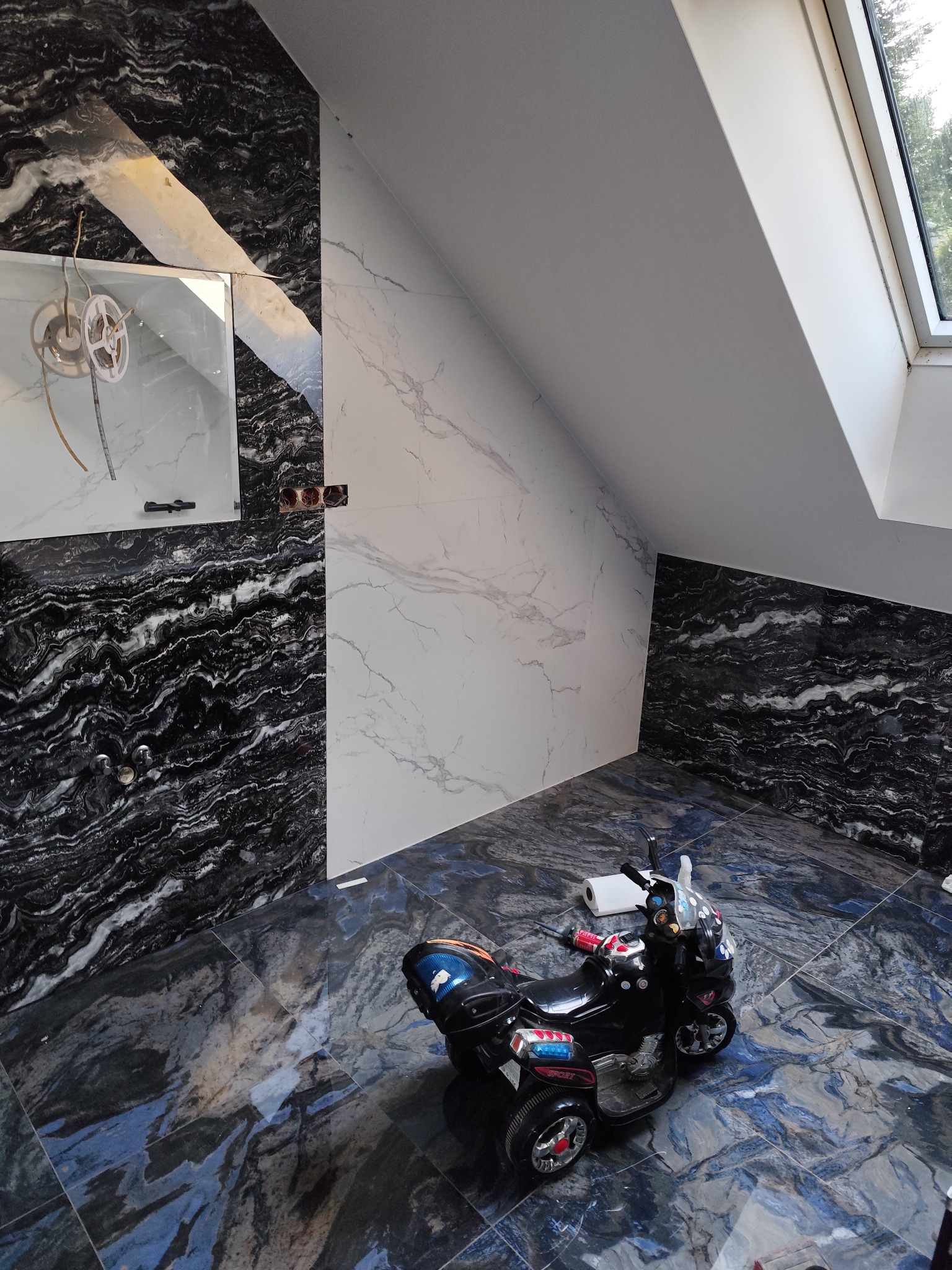 Wykończona łazienka na poddaszu z czarno-białymi marmurowymi płytkami, oknem dachowym i dziecięcym motorem na podłodze. Widoczne niedokończone podłączenie oświetlenia.