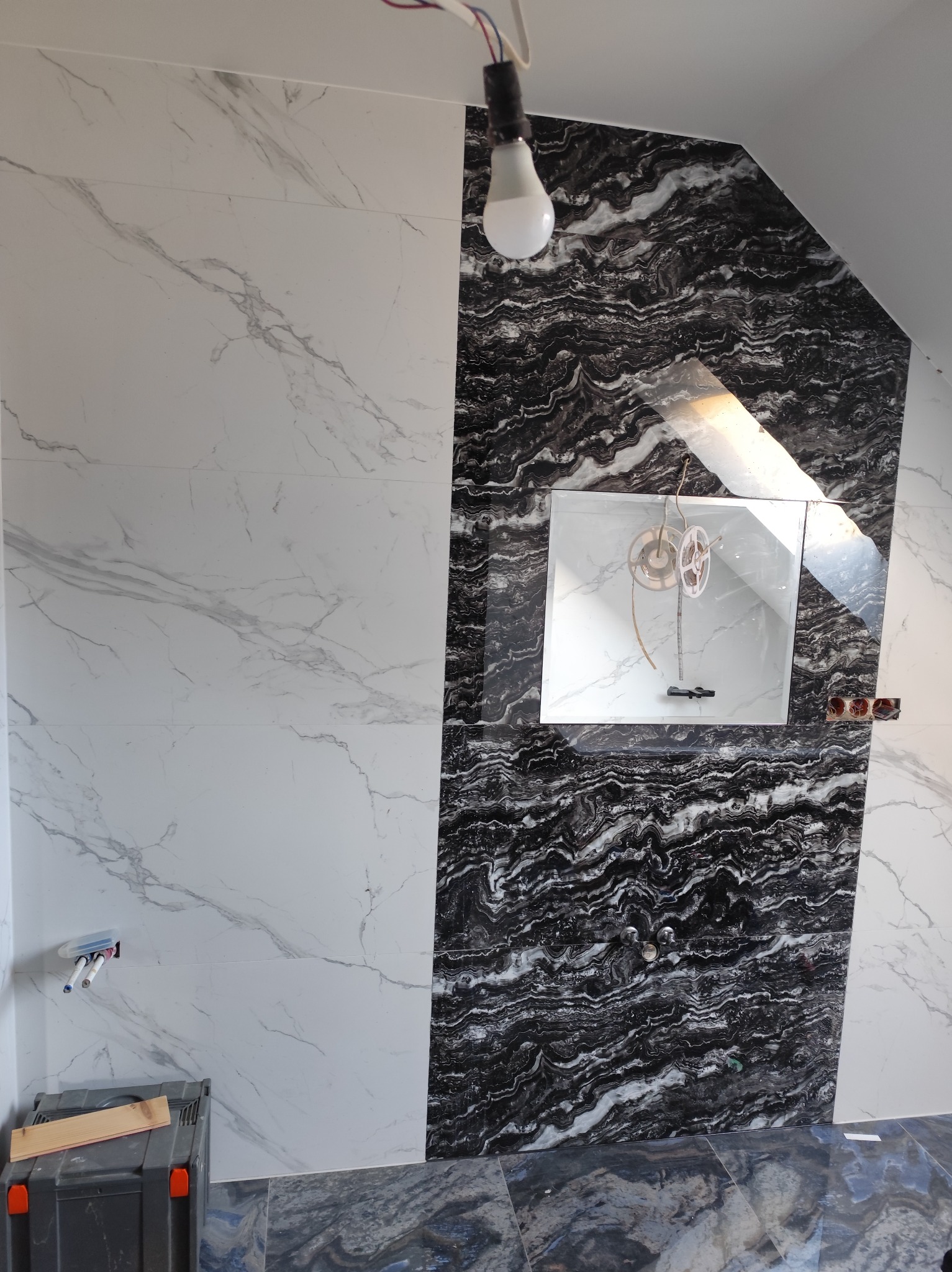 Wykończona ściana łazienki z białymi i czarnymi płytkami imitującymi marmur. Widoczne przygotowane przyłącza wodne i elektryczne. Na podłodze ułożone płytki.