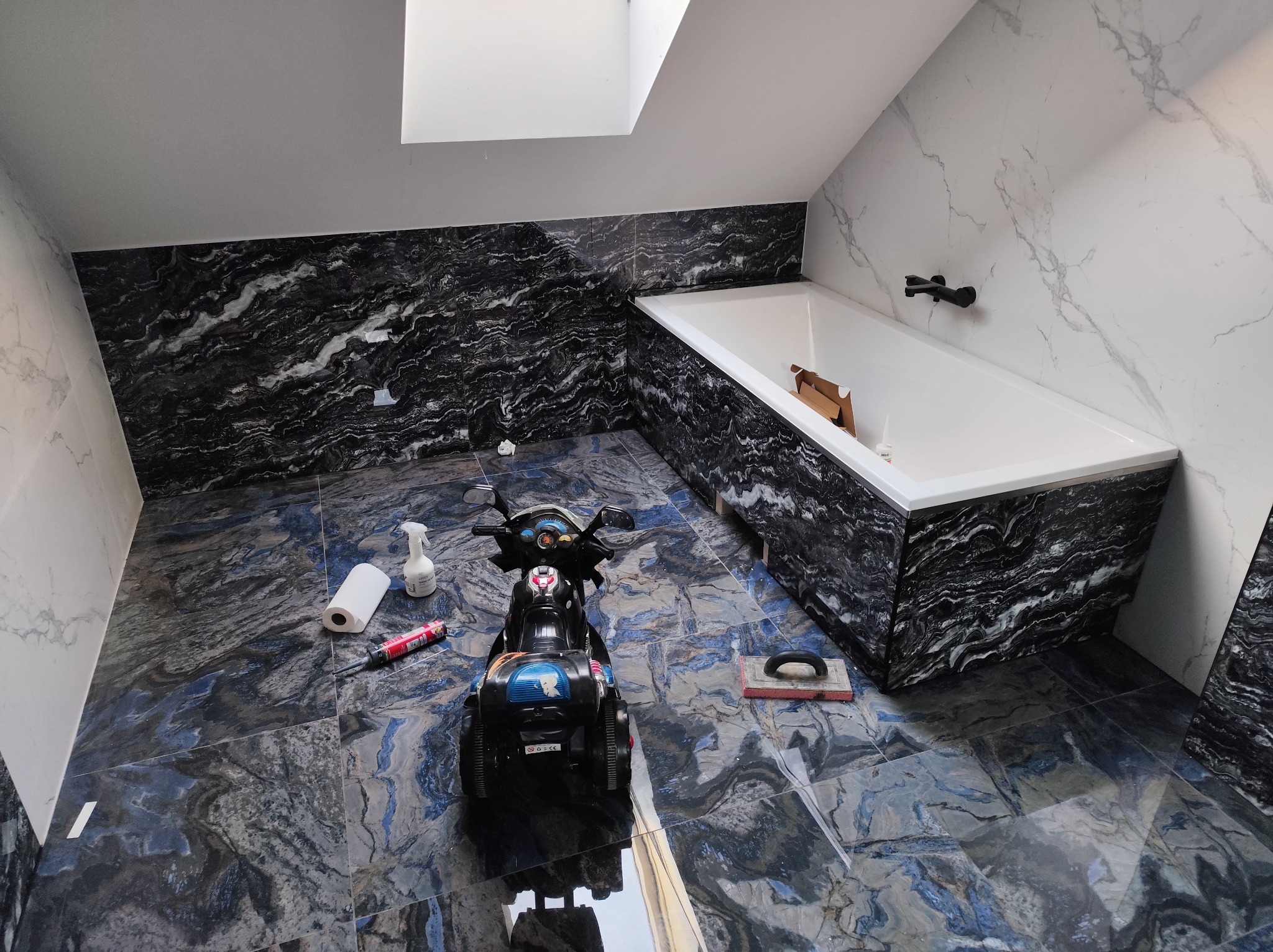 Wykończona łazienka na poddaszu z wanną obudowaną czarnym marmurem, niebieska podłoga z płytek, zabawkowy motor i narzędzia glazurnicze.