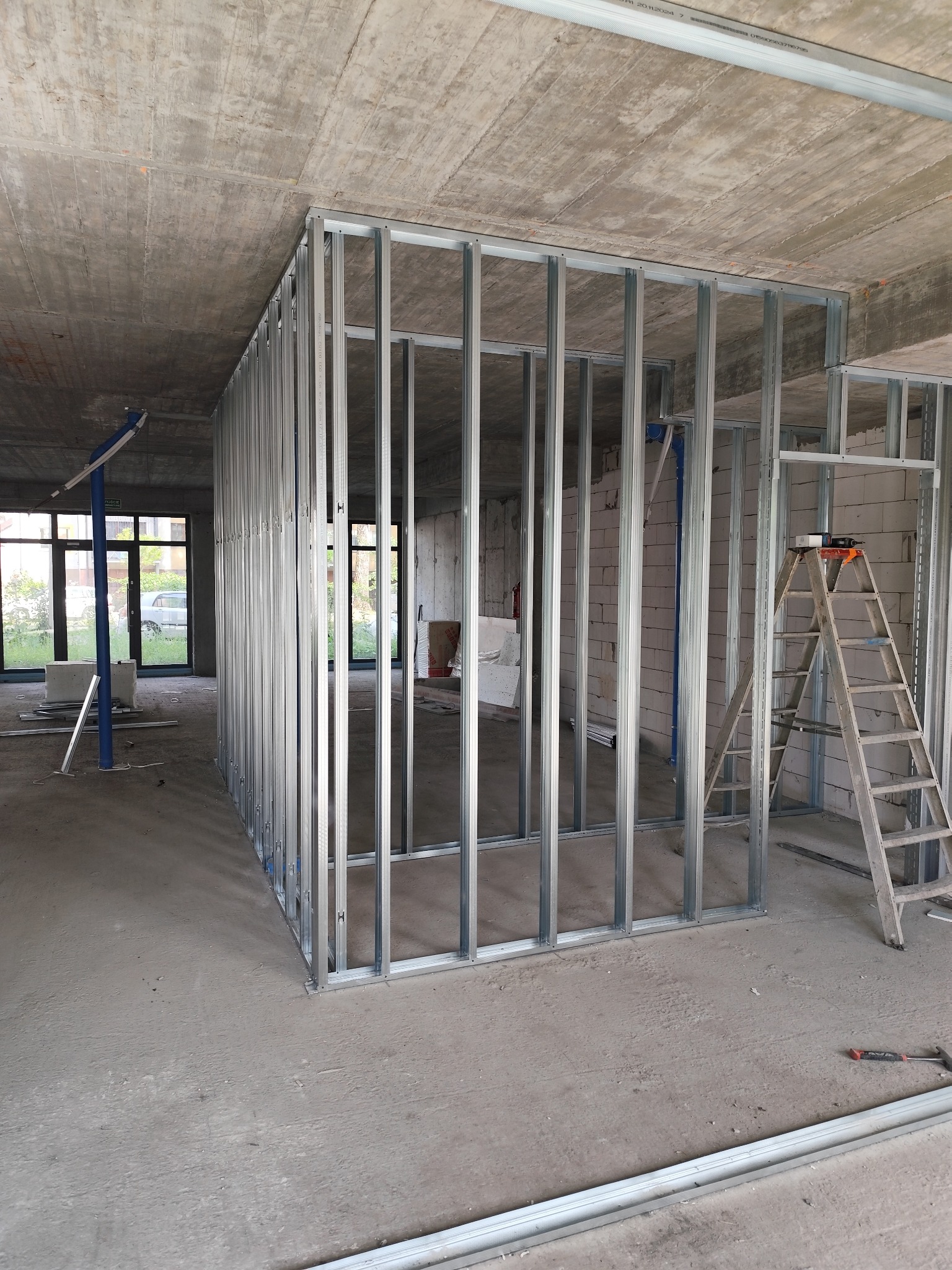 Konstrukcja ścianki działowej z profili stalowych na surowym betonie, widoczna drabina i narzędzia. W tle okna i materiały budowlane. Wnętrze w trakcie prac wykończeniowych.