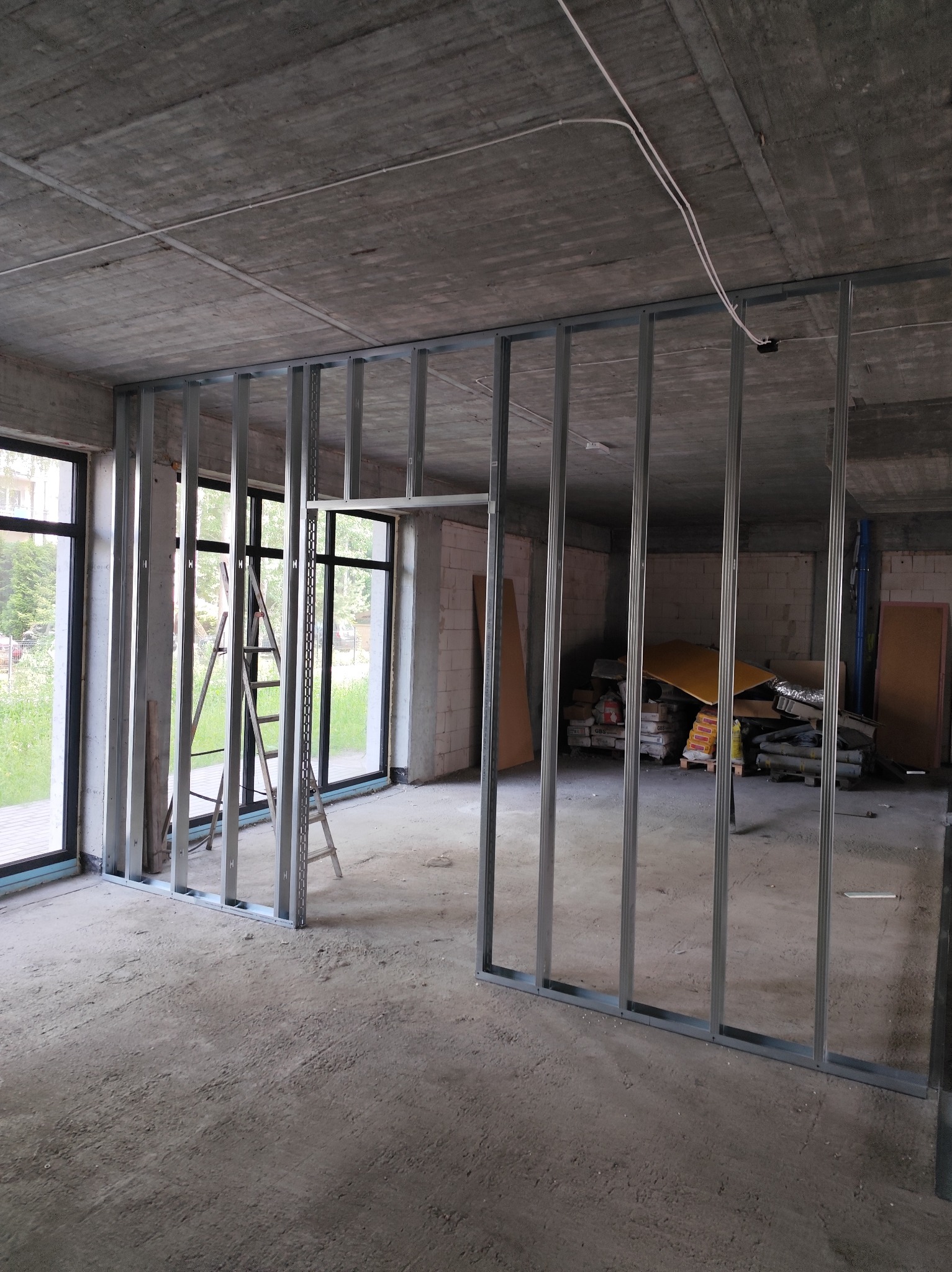Konstrukcja ścian działowych z profili stalowych w surowym wnętrzu. Widoczne okna, drabina i materiały budowlane. Szare, betonowe ściany i podłoga w trakcie prac.