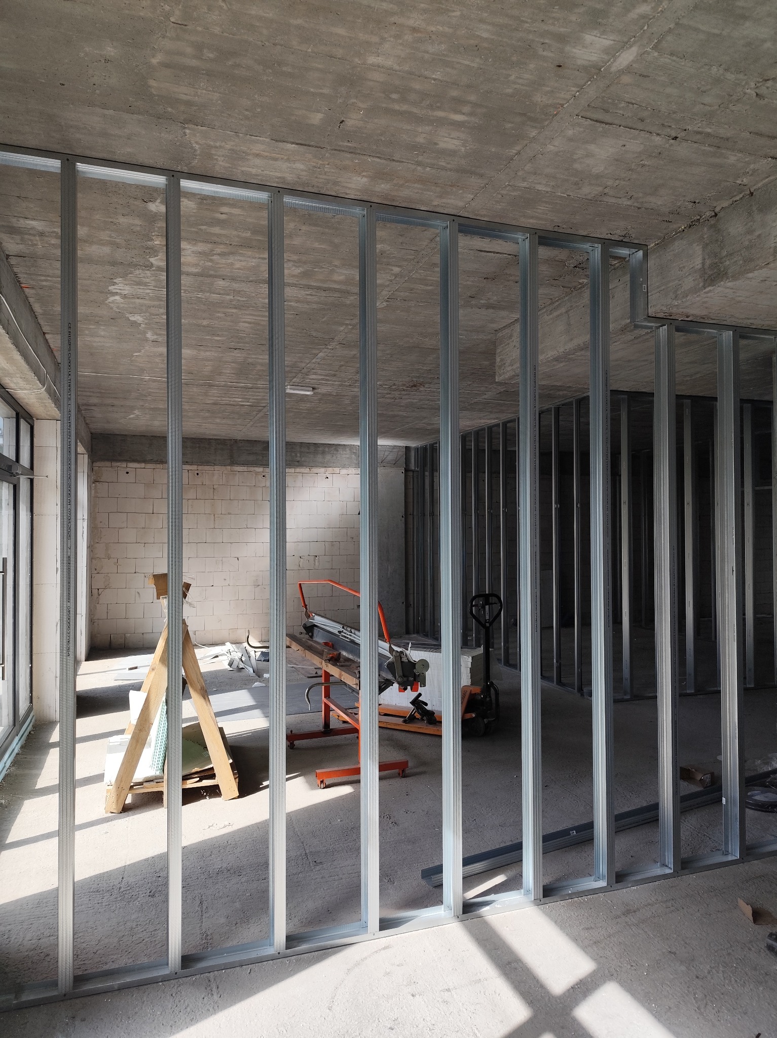 Montaż metalowej konstrukcji ścian działowych w surowym wnętrzu. Widoczne narzędzia i materiały budowlane. Betonowy sufit i ściany z białego pustaka.