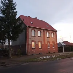 Dwupiętrowy dom z nowym, czerwonym dachem z dachówki, widoczny z poziomu ulicy, z wysoką choinką obok i fragmentem zaśnieżonego trawnika.