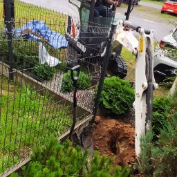 Firma Usługowo Handlowa Waldemar Słonina - Operator w minikoparce, wykop w ziemi blisko ogrodzenia. Prace ziemne na posesji z zielenią. Widoczna łopata i fragment niebieskiej plandeki. Dzień pochmurny.