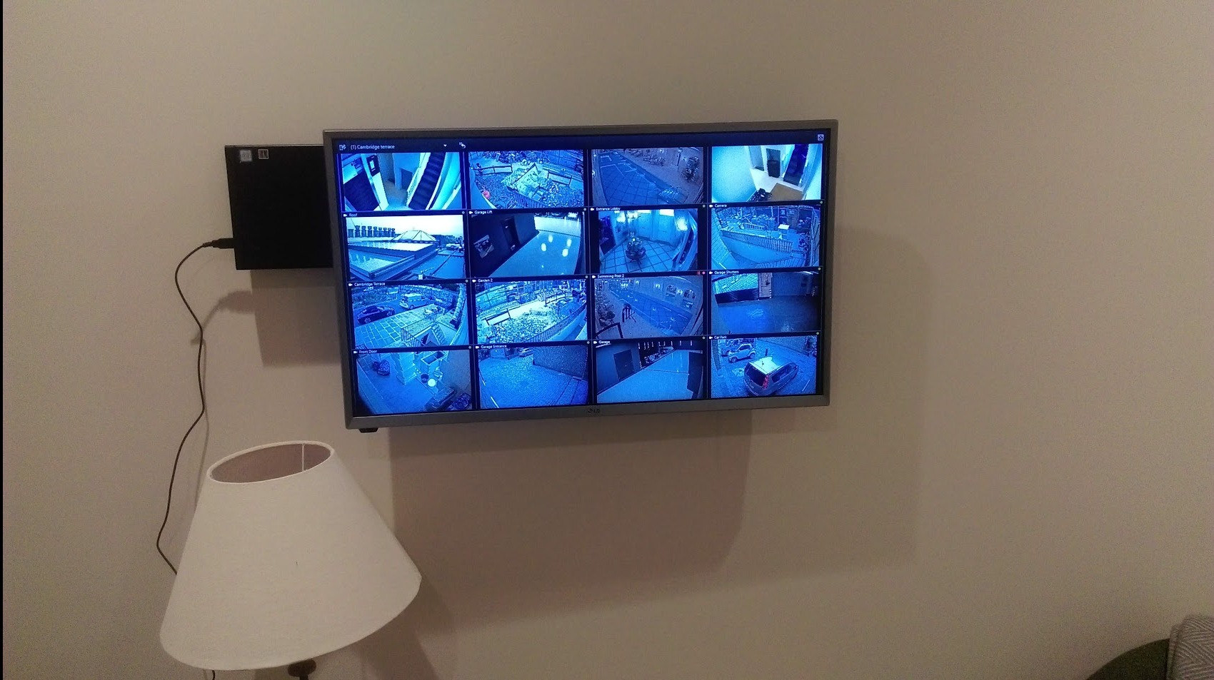 Ekran monitora LCD wyświetlający podgląd z wielu kamer bezpieczeństwa, zamontowany na ścianie obok lampy stojącej, z widocznym rejestratorem DVR.