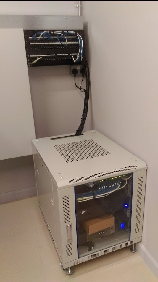 Szafa rack z osprzętem sieciowym, widoczne kable, patch panel i zasilacz UPS Eaton, umieszczona w rogu pomieszczenia.
