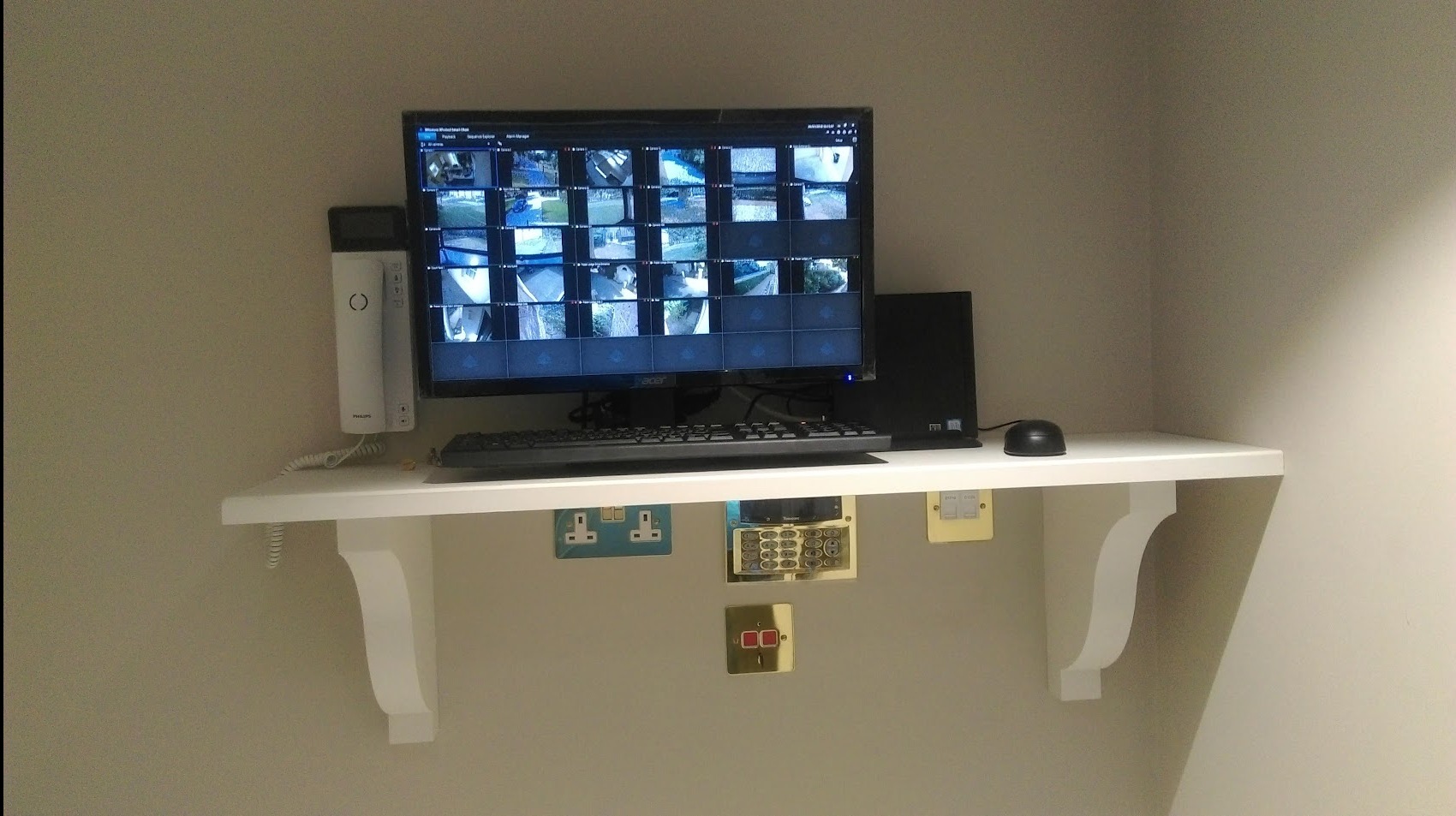 Monitor wyświetlający obraz z wielu kamer monitoringu, umieszczony na białej półce wspartej na ozdobnych wspornikach, obok domofonu Philips, pod półką widoczne gniazdka elektryczne i klawiatura...