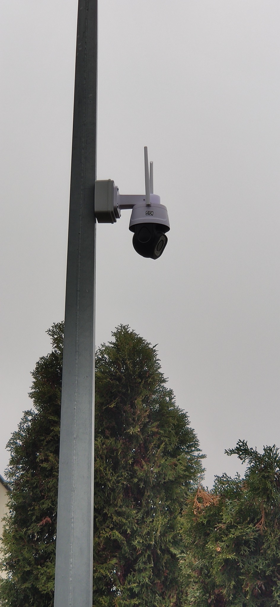 Zewnętrzna kamera monitoringu 4K z antenami, zamontowana na metalowym słupie, na tle zielonych drzew i pochmurnego nieba.