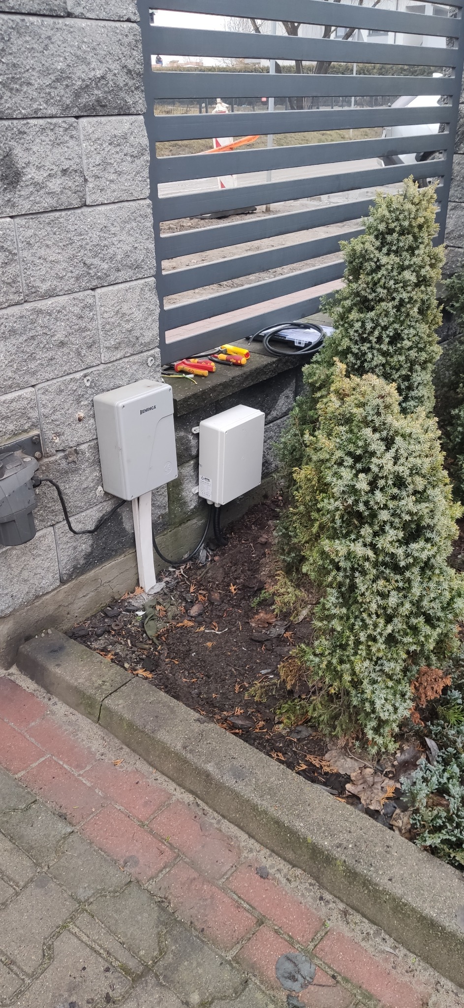 Dwa szare moduły sterujące, jeden z logo BENINCA, zamontowane na szarym murze obok szarej bramy posesji; obok leżą narzędzia i kable.