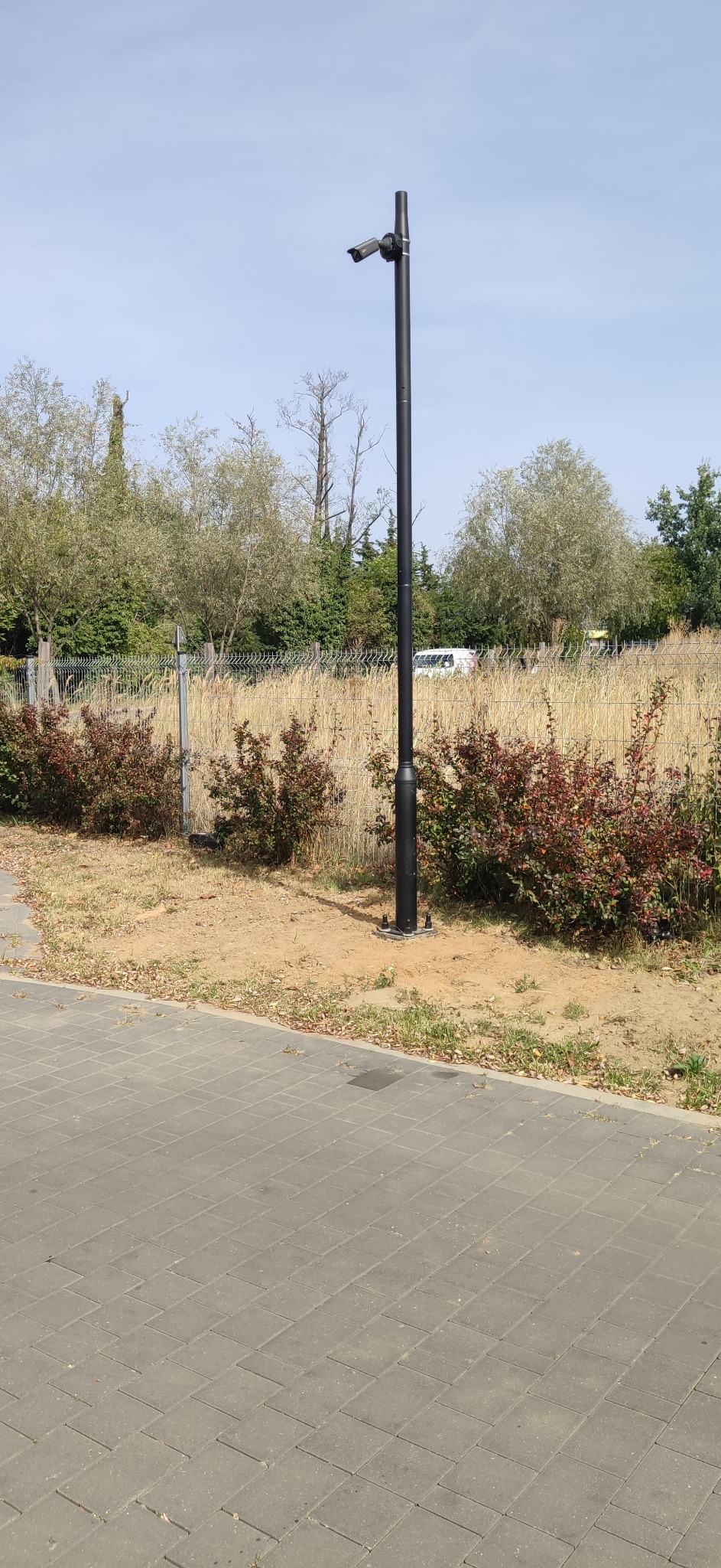 Kamera przemysłowa na czarnym słupie, zainstalowana na zewnątrz, z widocznymi krzewami i drzewami w tle. Jasne, dzienne światło.