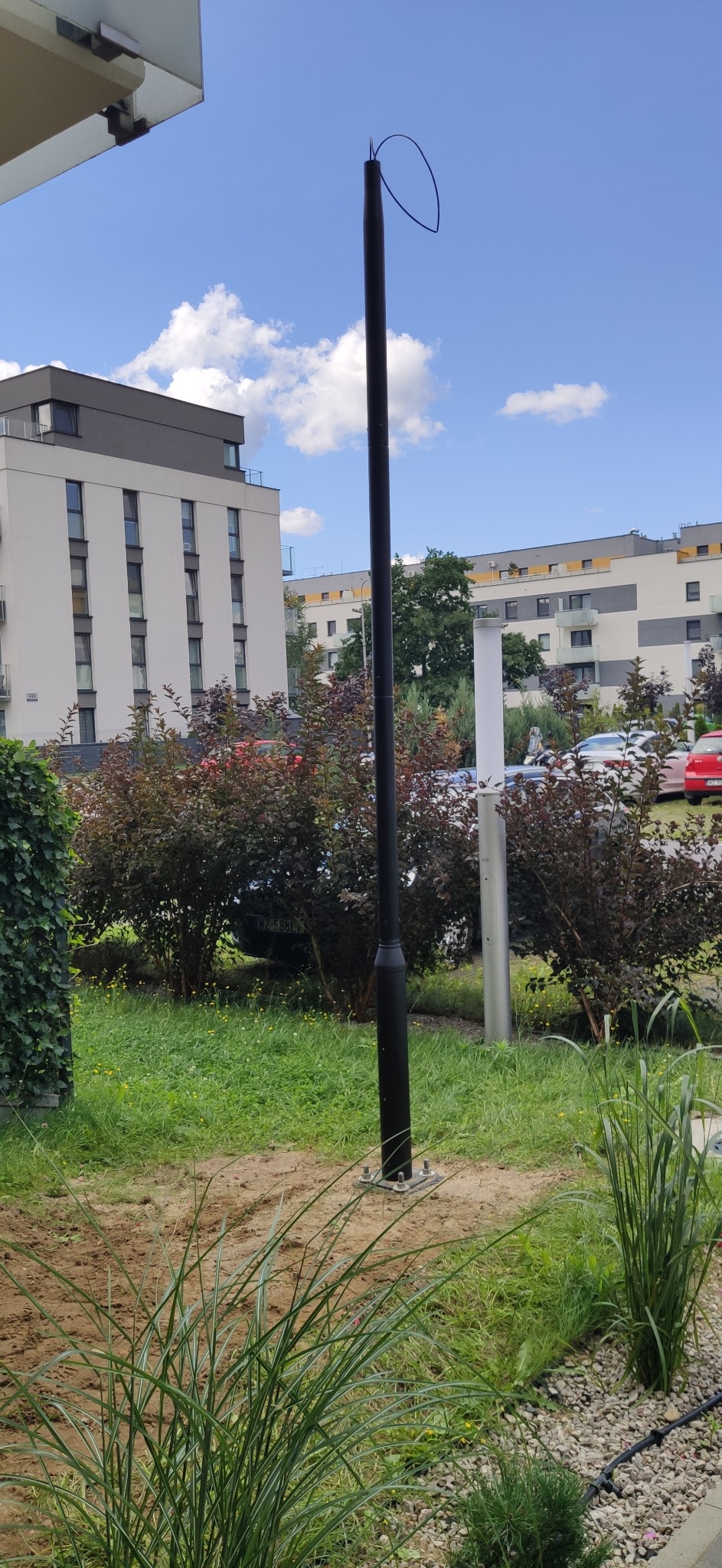 Czarny słup oświetleniowy bez lampy, wystający z ziemi na osiedlu. Widoczne przewody na górze słupa, budynki w tle i zieleń. Słoneczny dzień.