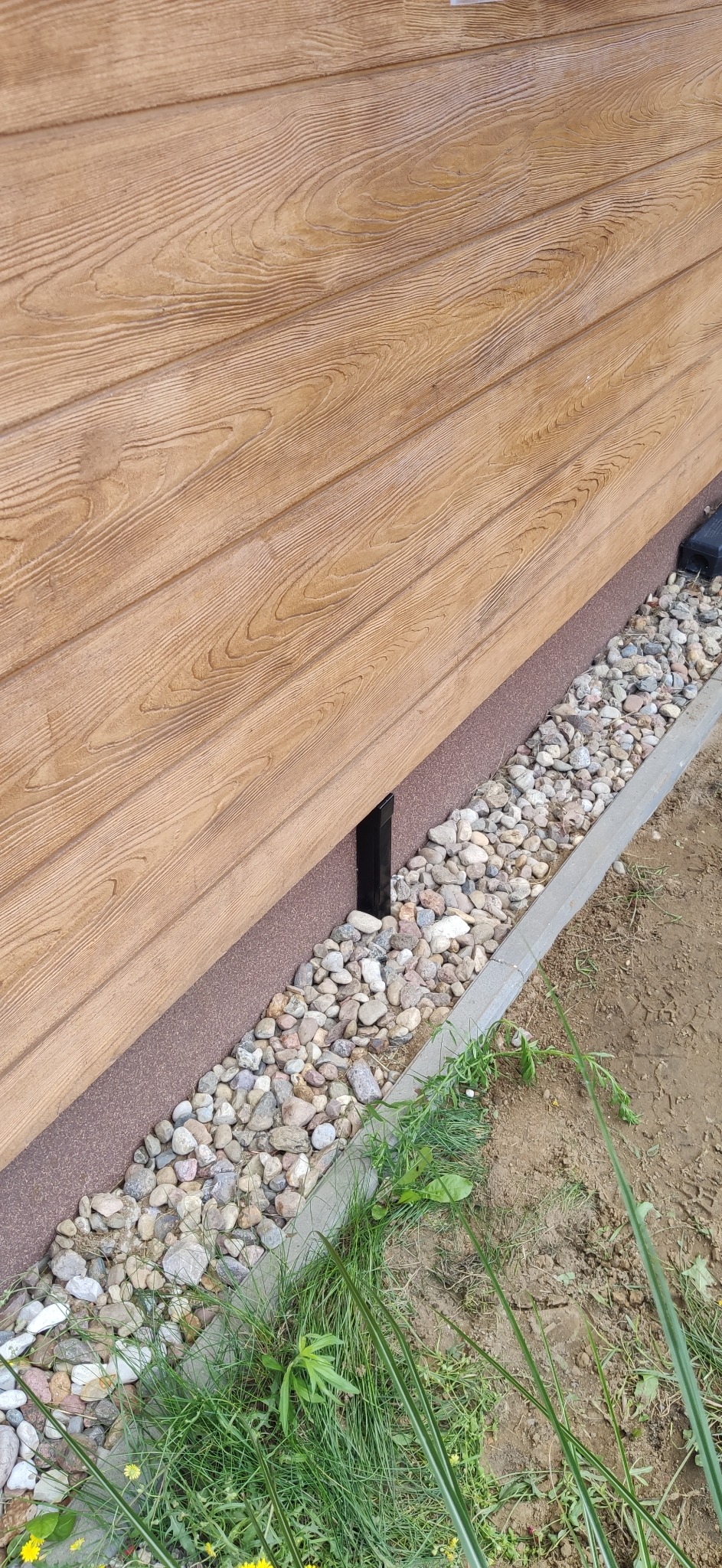 Fragment elewacji domu z drewnianą okładziną i kamiennym obrzeżem. Widoczna czarna rura i roślinność przy gruncie. Ujęcie z bliska, naturalne oświetlenie.