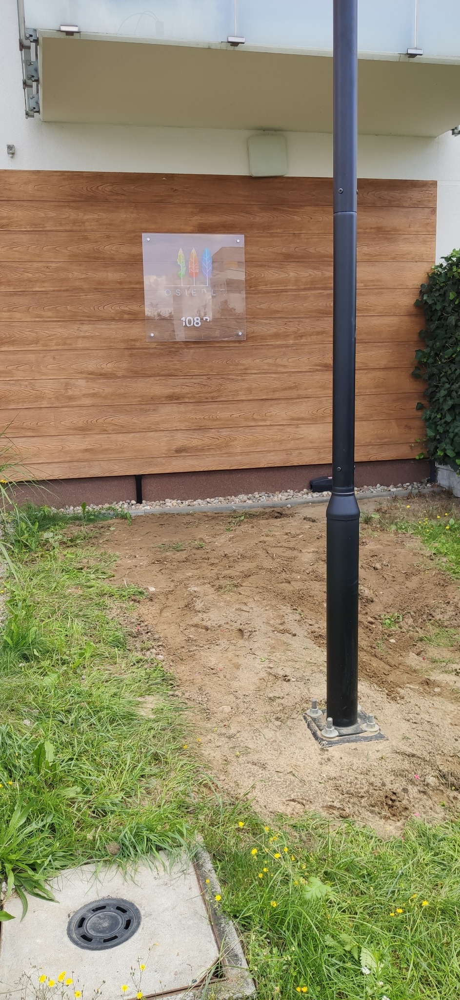 Czarna lampa ogrodowa, zamontowana na ziemi przy budynku z drewnianą elewacją i tabliczką z numerem 108, otoczona trawą i piaskiem.
