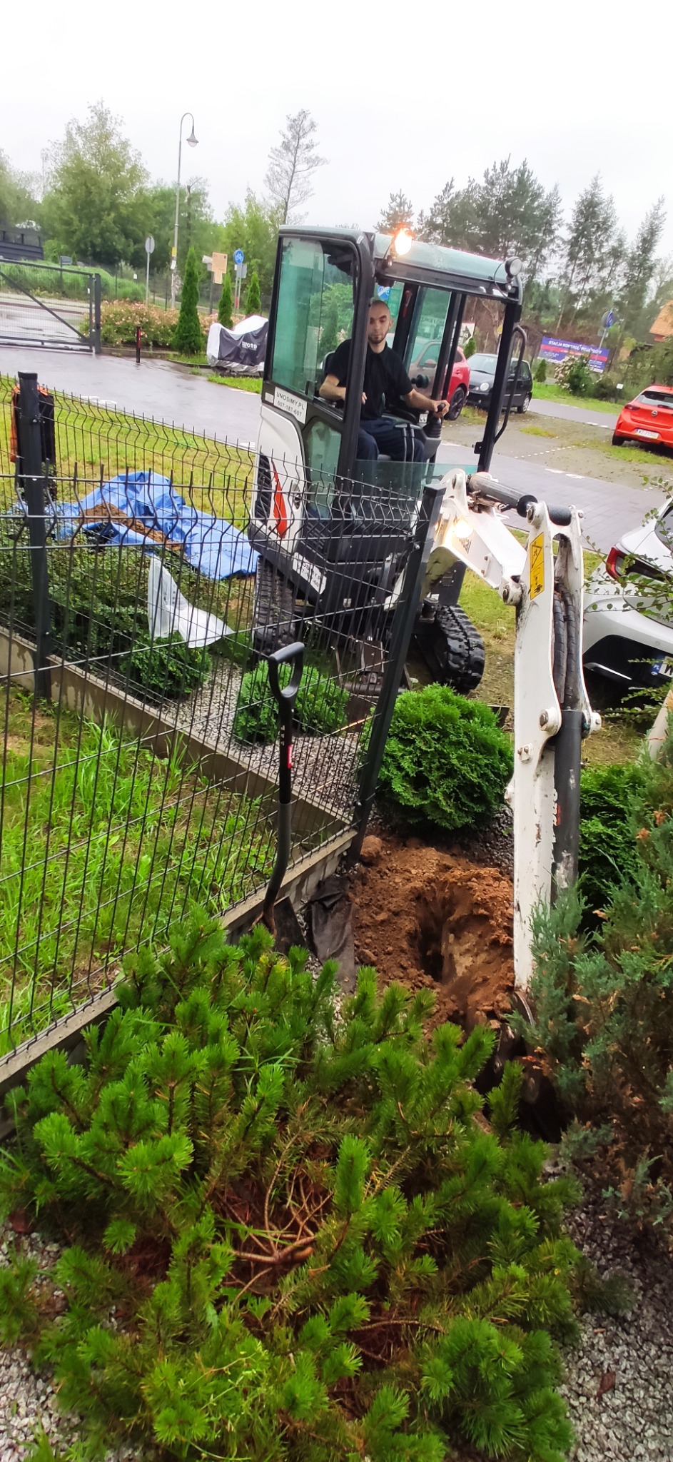 Operator w minikoparce, wykop w ziemi blisko ogrodzenia. Prace ziemne na posesji z zielenią. Widoczna łopata i fragment niebieskiej plandeki. Dzień pochmurny.