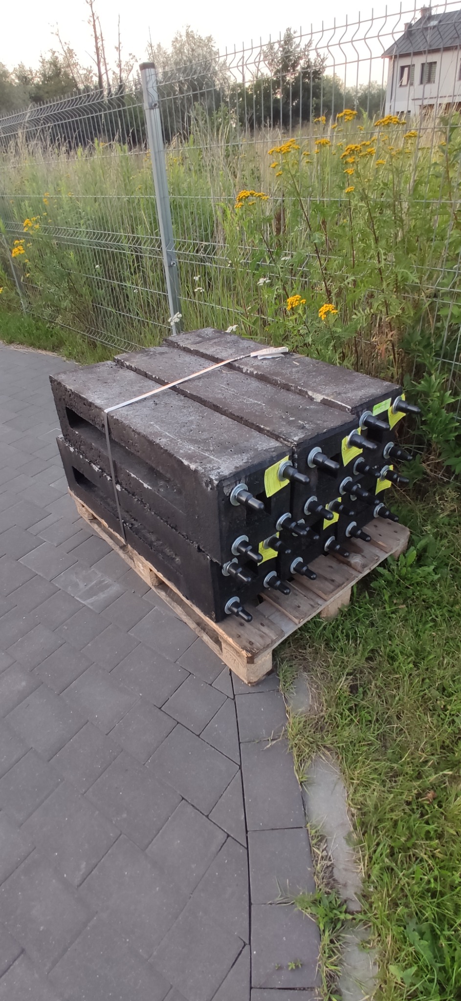 Betonowe prefabrykaty z metalowymi elementami mocującymi, ułożone na palecie, obok metalowego ogrodzenia i kostki brukowej. Widoczne żółte kwiaty na tle ogrodzenia.