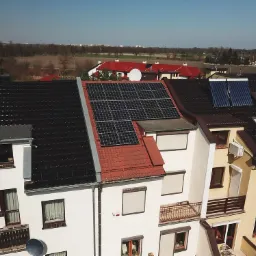 Widok z góry na szereg domów z zainstalowanymi panelami słonecznymi na dachach, w tle widoczny krajobraz podmiejski.