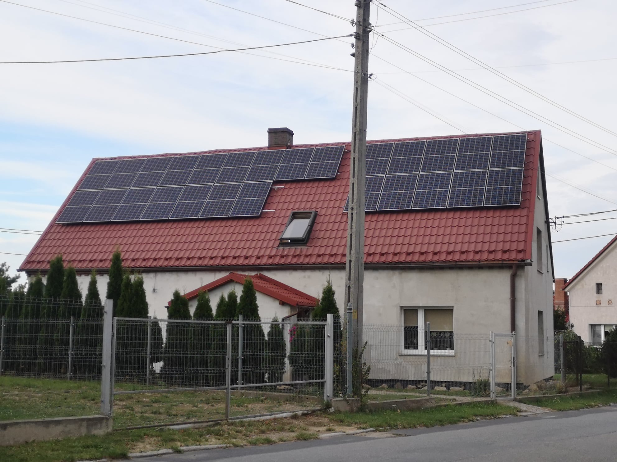 Dom jednorodzinny z panelami fotowoltaicznymi na czerwonym dachu, widok zza metalowego ogrodzenia, słup elektryczny po prawej stronie.