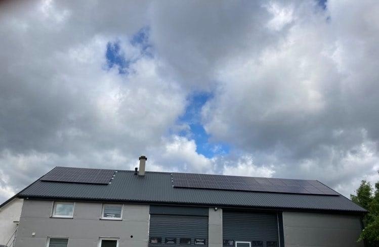 Panele słoneczne zamontowane na dachu budynku magazynowego z szarą elewacją i bramami rolowanymi, widoczne na tle pochmurnego nieba.