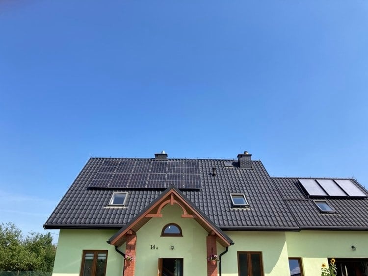 Dom jednorodzinny z zainstalowanymi panelami słonecznymi na dachu, widok z dołu, błękitne niebo w tle.