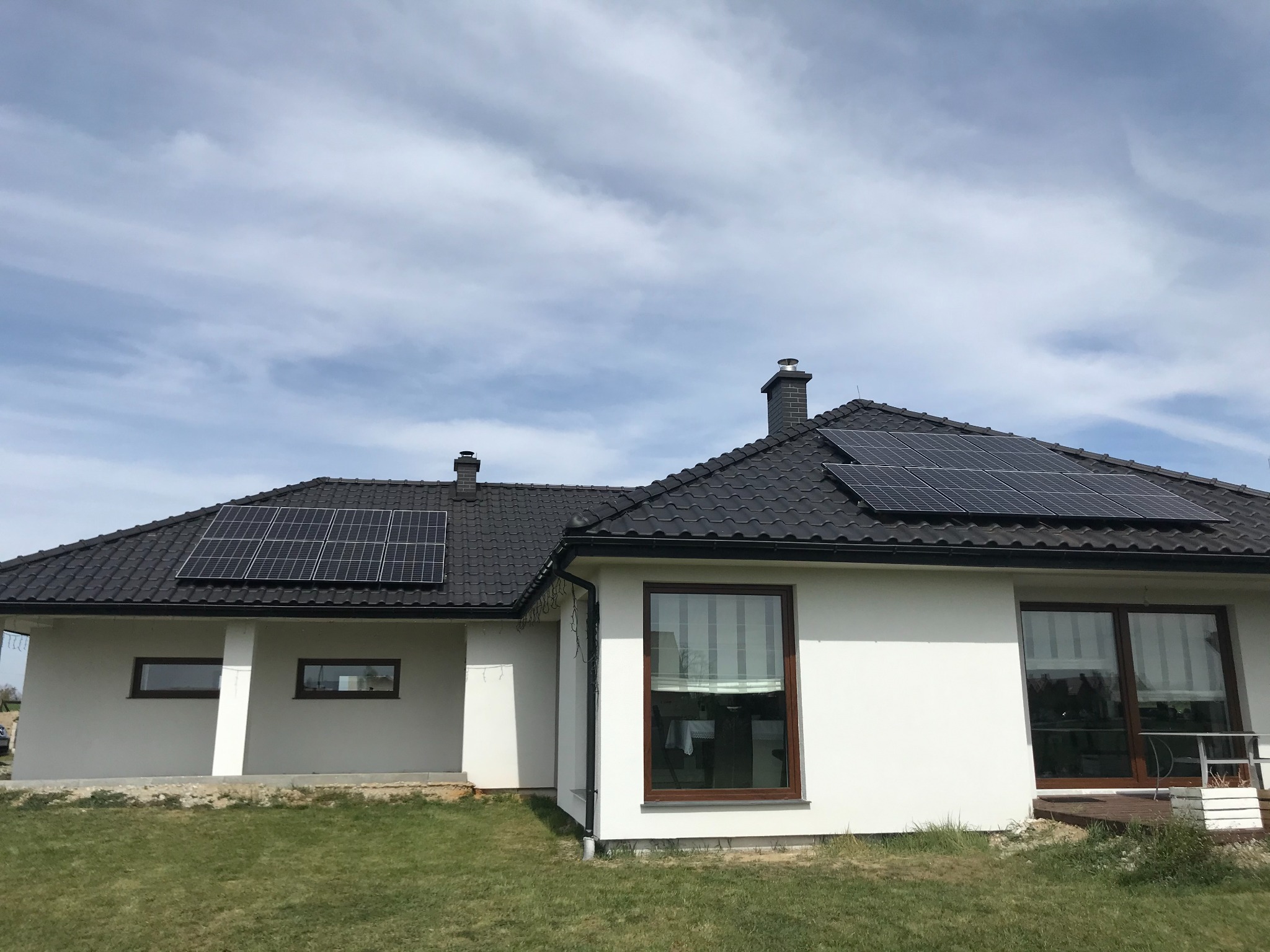 Nowoczesny dom jednorodzinny z ciemną dachówką i panelami słonecznymi na dachu, widok z poziomu trawnika w słoneczny dzień.