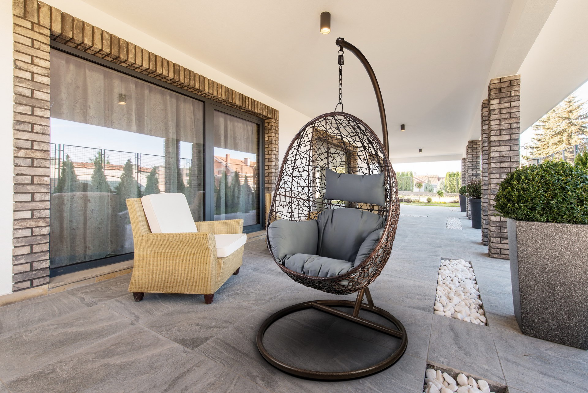 Zadaszony taras z szarymi płytkami, wiklinowym fotelem i wiszącym fotelem-kokon z szarymi poduszkami, dekorowany ceglanymi filarami i donicami z zielonymi krzewami.