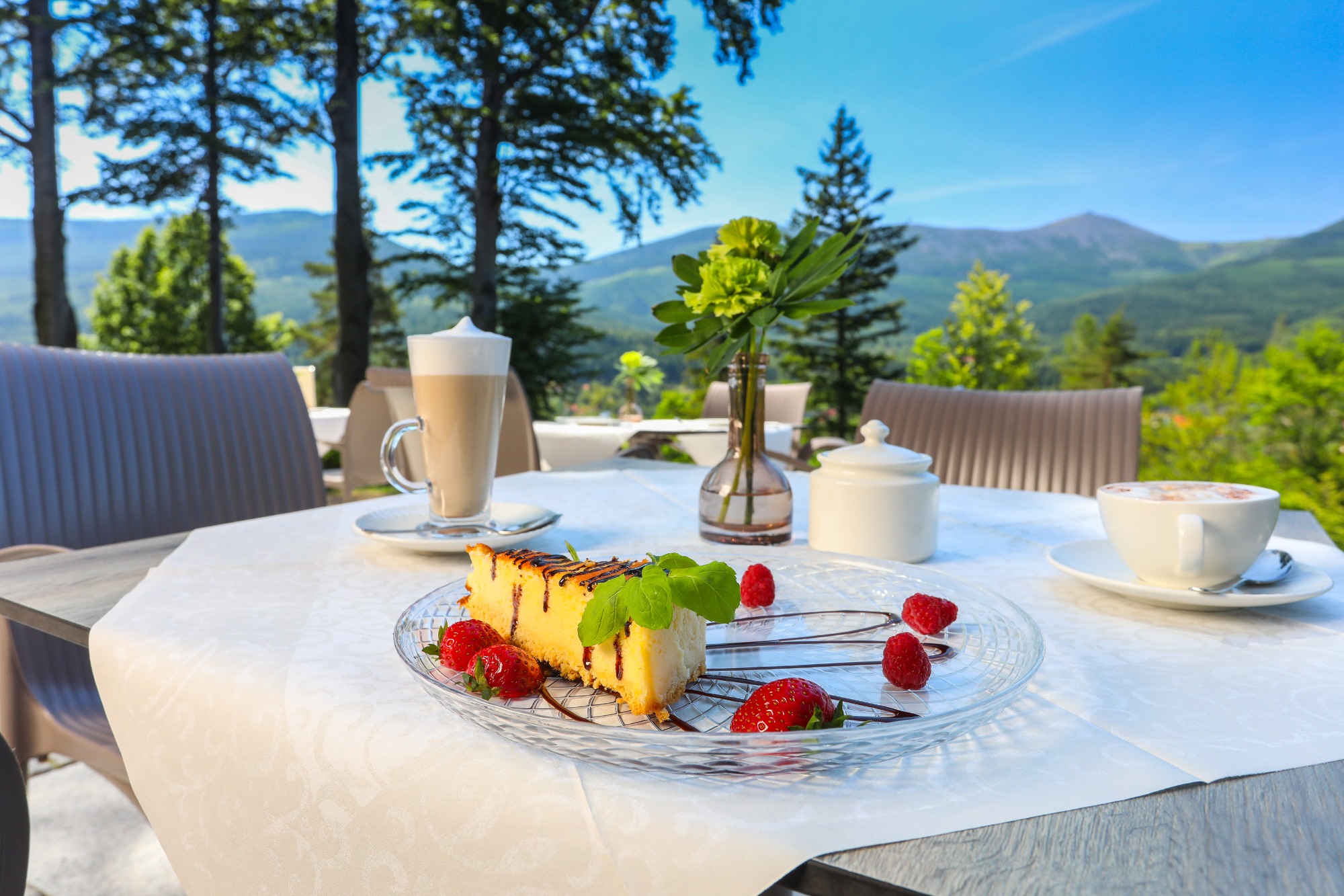 Kawa latte w szklance, cappuccino w filiżance, sernik polany czekoladą z truskawkami i malinami na szklanym talerzu, wazon z kwiatami na stole z białym obrusem, w tle widok na góry i las.