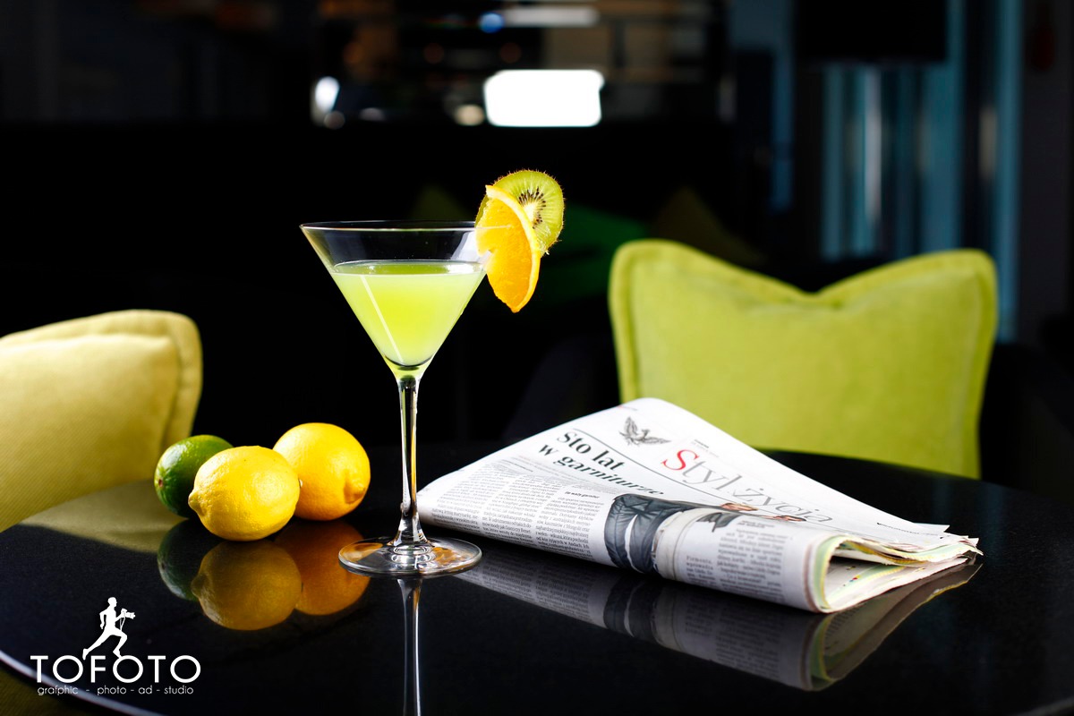 Martini z plasterkiem pomarańczy i kiwi, cytryny i gazeta na czarnym, błyszczącym stole z odbiciami, w tle zielone poduszki.