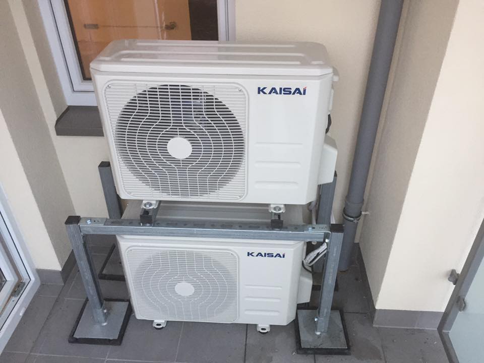 Dwa białe agregaty klimatyzacyjne marki Kaisai, zamontowane jeden nad drugim na metalowej konstrukcji wsporczej, umieszczone na zewnątrz budynku przy ścianie i rurze spustowej.