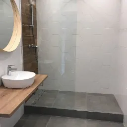 Nowoczesna łazienka z kabiną prysznicową typu walk-in, heksagonalnymi białymi płytkami na ścianie, drewnianym blatem pod umywalką i okrągłym lustrem.