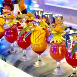 Rząd kolorowych koktajli w kieliszkach, ozdobionych egzotycznymi owocami, ustawionych na blacie z efektem suchego lodu, w tle barowe akcesoria.