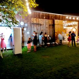 Dobrze.Pstryknięci - Grupa osób pozuje do zdjęcia w fotobudce na tle drewnianej stodoły udekorowanej światełkami i ozdobami.