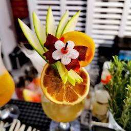 Dobrze.Pstryknięci - Dekoracyjny drink z suszoną pomarańczą, kwiatem z wiśnią i listkami ogórka na barowym blacie z logo Jack Daniel's, obok doniczka z ziołami i inny drink.