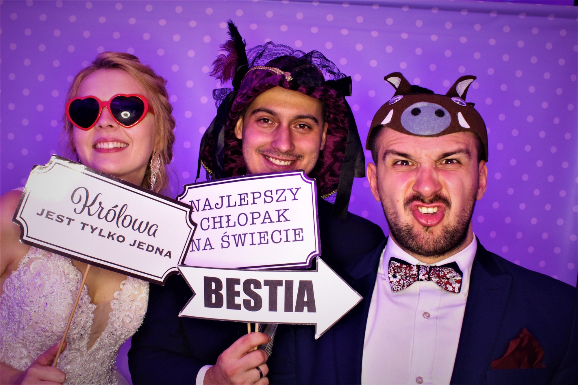 Trzy osoby pozują w fotobudce w Chrzęsnem, trzymając rekwizyty z napisami: 'Królowa jest tylko jedna', 'Najlepszy chłopak na świecie' i strzałka z napisem 'Bestia'. Kobieta w sukni i okularach...