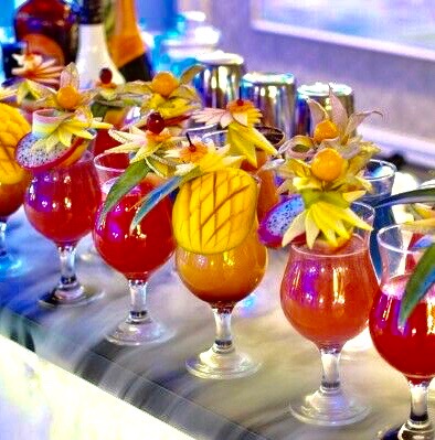 Rząd kolorowych koktajli w kieliszkach, ozdobionych egzotycznymi owocami, ustawionych na blacie z efektem suchego lodu, w tle barowe akcesoria.