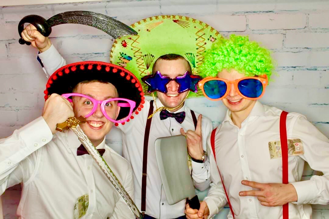 Trzech mężczyzn w przebraniach pozuje do zdjęcia w fotobudce: jeden w sombrero i różowych okularach trzymający plastikowy miecz, drugi w gigantycznych okularach w kształcie gwiazd i z tasakiem...