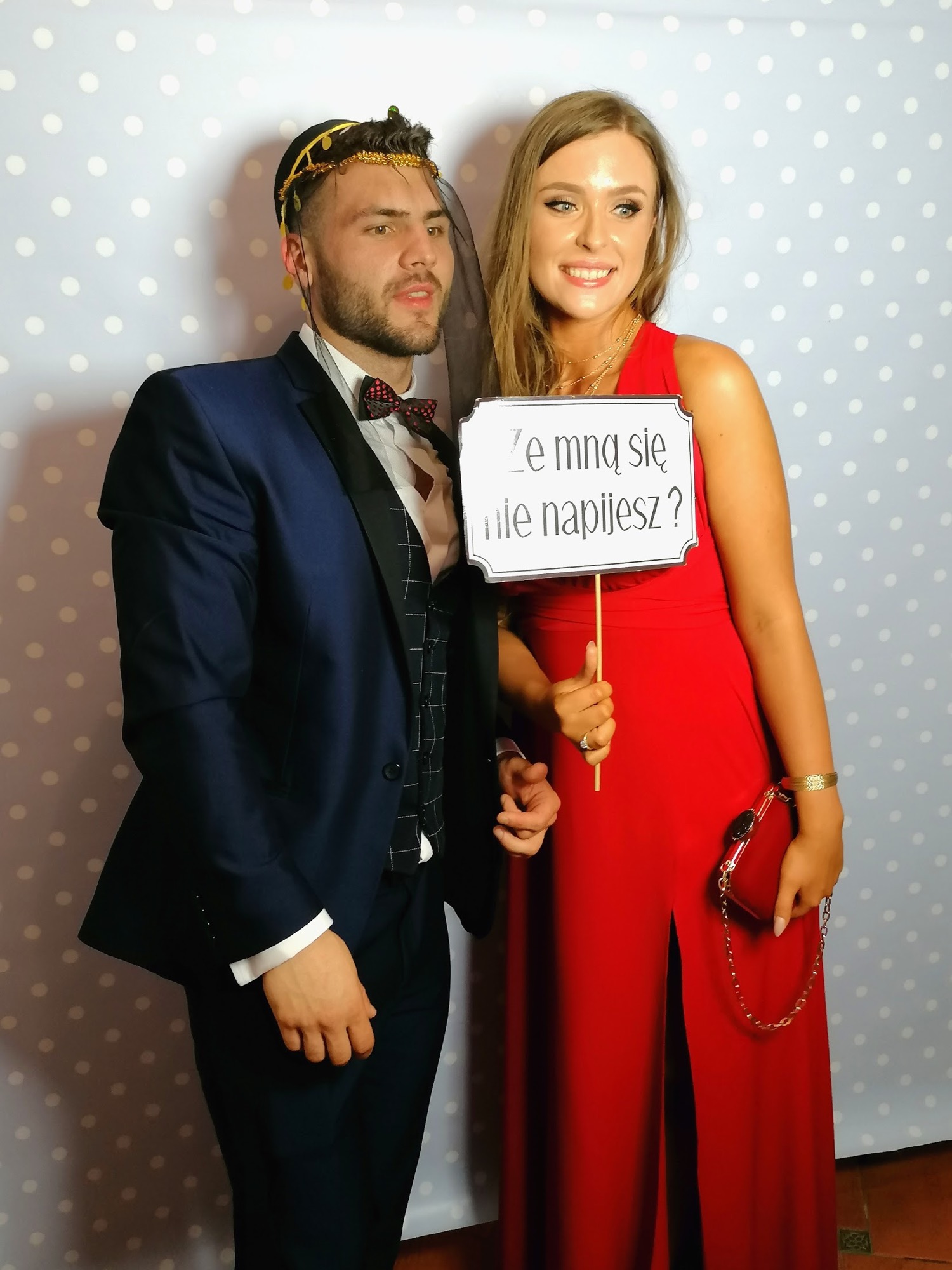 Para pozuje ze śmiesznym rekwizytem z napisem 'Ze mną się nie napijesz?' na tle w kropki. Mężczyzna w granatowym garniturze i muszce, kobieta w długiej czerwonej sukni z rozcięciem.