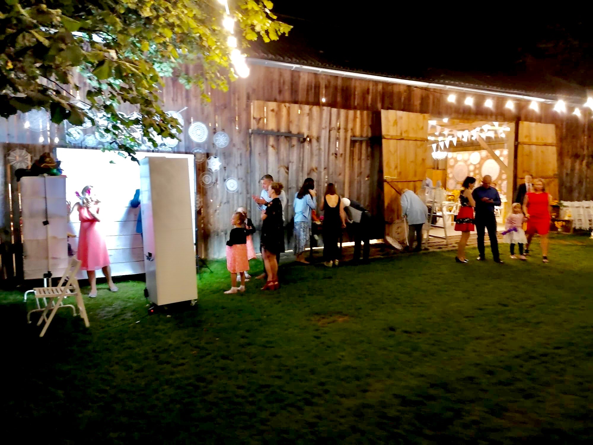 Grupa osób pozuje do zdjęcia w fotobudce na tle drewnianej stodoły udekorowanej światełkami i ozdobami.