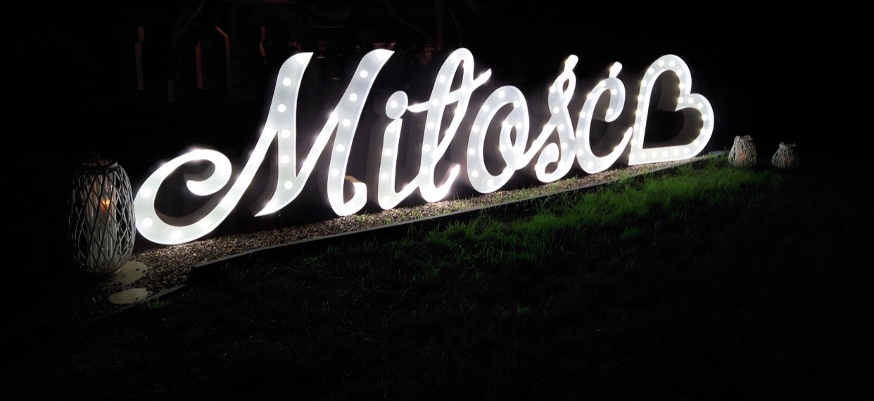 Podświetlany napis 'Miłość' z żarówkami, umieszczony na trawie, obok lampion i dekoracyjne kule, nocna sceneria.