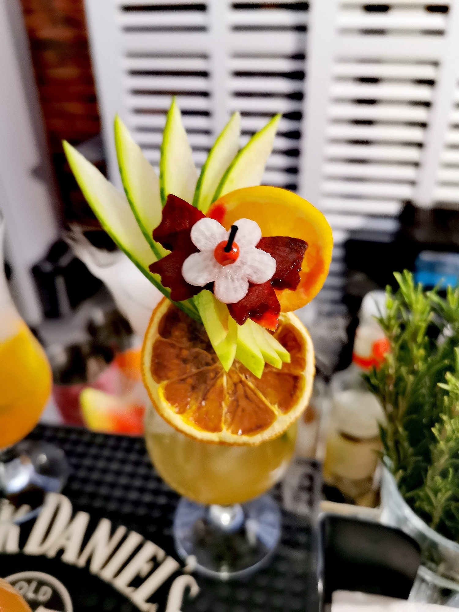 Dekoracyjny drink z suszoną pomarańczą, kwiatem z wiśnią i listkami ogórka na barowym blacie z logo Jack Daniel's, obok doniczka z ziołami i inny drink.