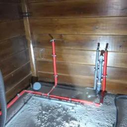 Instalacja hydrauliczna z czerwonych i szarych rur w drewnianym domu w trakcie montażu, widoczne połączenia zaciskowe.