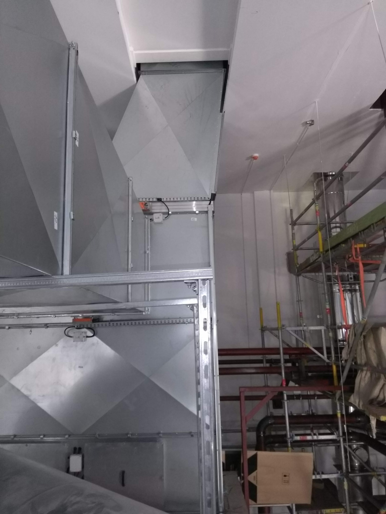 Widok z dołu na montaż dużego, metalowego kanału wentylacyjnego w budynku, z widocznymi elementami mocującymi, przewodami i rusztowaniem w tle.